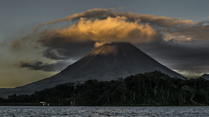 Volcano with ominous cloud cover at sunset over dark forest and water, evoking creepy facts about the world atmosphere.