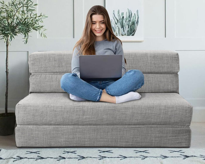 Young woman sitting cross-legged on a small gray ANONER couch, working on a laptop in a stylish living space.