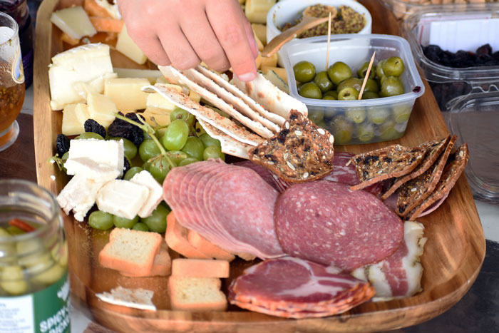 Hand reaching for crackers on a $70 charcuterie board with meats, cheeses, grapes, olives, and crackers at Thanksgiving. Hand reaching for crackers on a $70 charcuterie board with meats, cheeses, grapes, olives, and crackers at Thanksgiving.