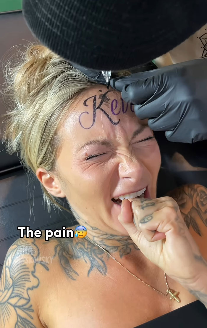 Influencer getting fake tattoo of boyfriend's name on forehead, wincing in pain, tattoo artist in black gloves.