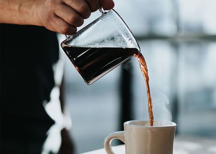Hand pouring coffee from glass carafe into white mug steaming in minimalist kitchen showcasing frugal tips concept.