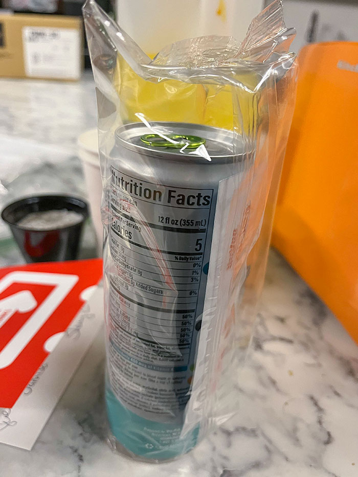 Slim can awkwardly packaged inside oversized plastic wrap, illustrating a facepalm-worthy packaging fail on a cluttered surface.