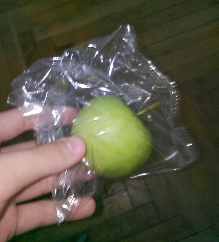 Hand holding a single green apple wrapped individually in unnecessary plastic packaging, showcasing packaging fails.