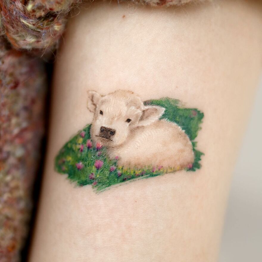 Tattoo of a calf resting on grass with flowers, showcasing nature and animal elements by a Korean ink artist.