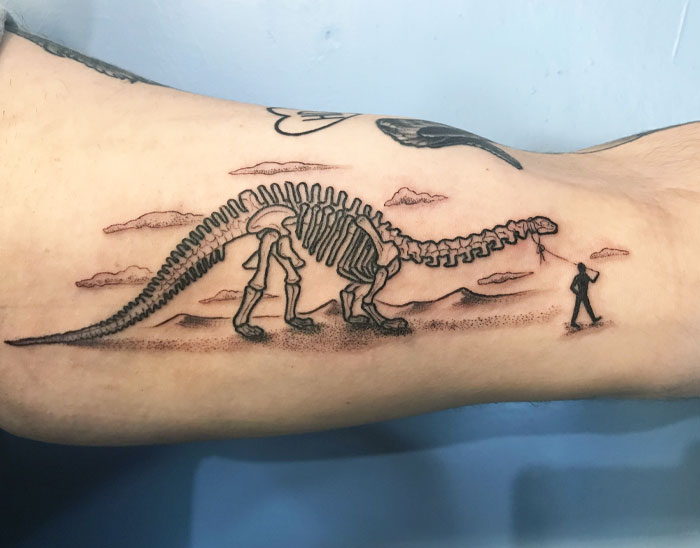 Dinosaur skeleton tattoo with person holding a leash, set against a desert landscape.