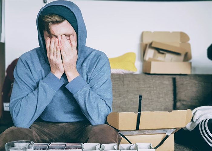Person in a blue hoodie sitting on a couch, covering their face, surrounded by open boxes, depicting burnout.