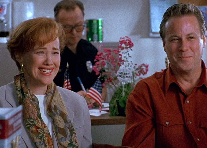 Catherine O'Hara from "Home Alone," smiling in a festive setting with flowers and flags, wearing a scarf and blazer.