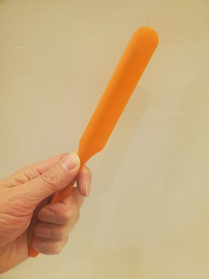 A hand holding a funny orange spatula-shaped product against a plain background.