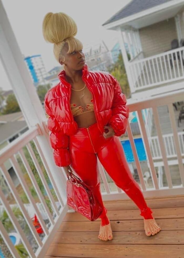 Person wearing a high bun, a red puffy jacket, and red pants with funny feet shoes on a balcony.
