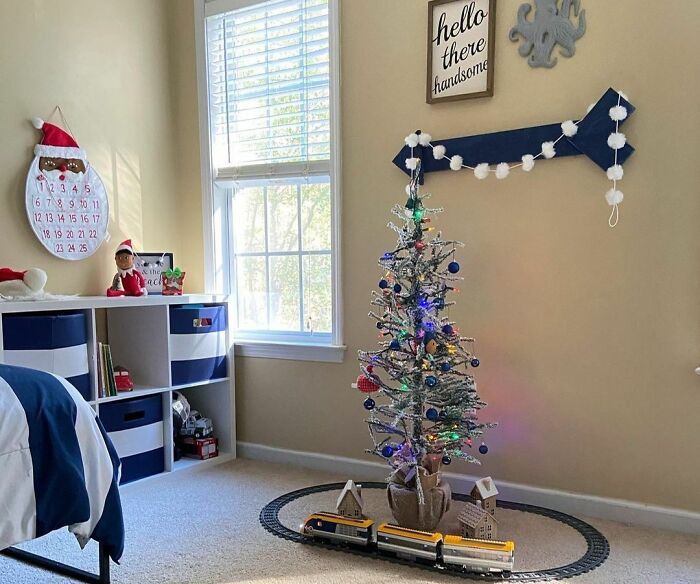 Small Christmas tree in kids room decorated affordably with train set and simple holiday ornaments near window.