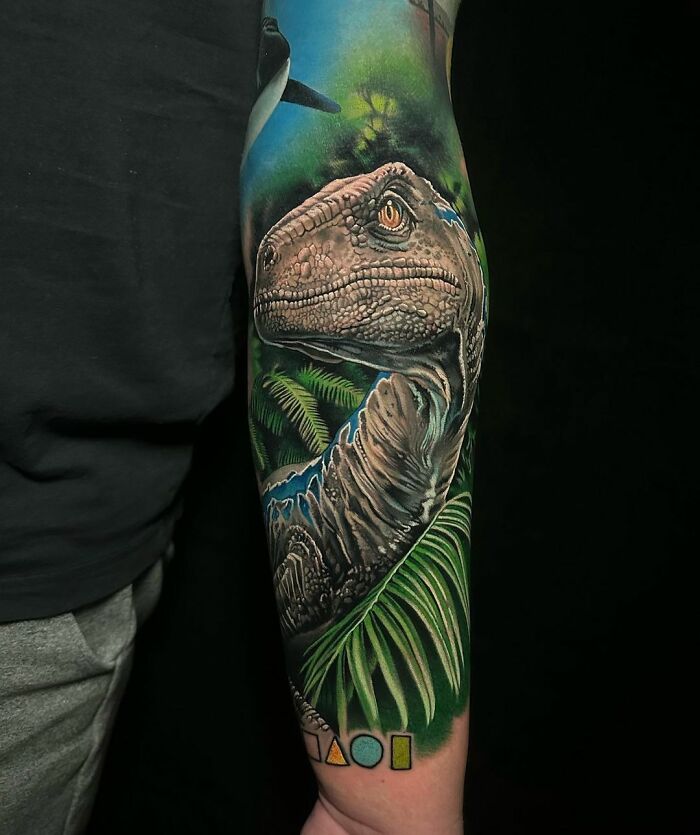 Cool dinosaur tattoo featuring a detailed Velociraptor surrounded by jungle elements on a person's arm.