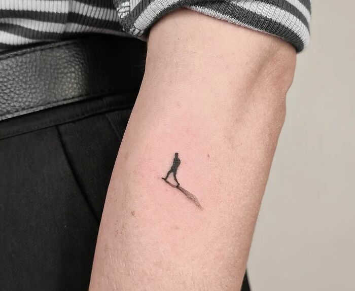 3D tattoo of a tiny walking figure on an arm, showcasing artistic shadow effect.