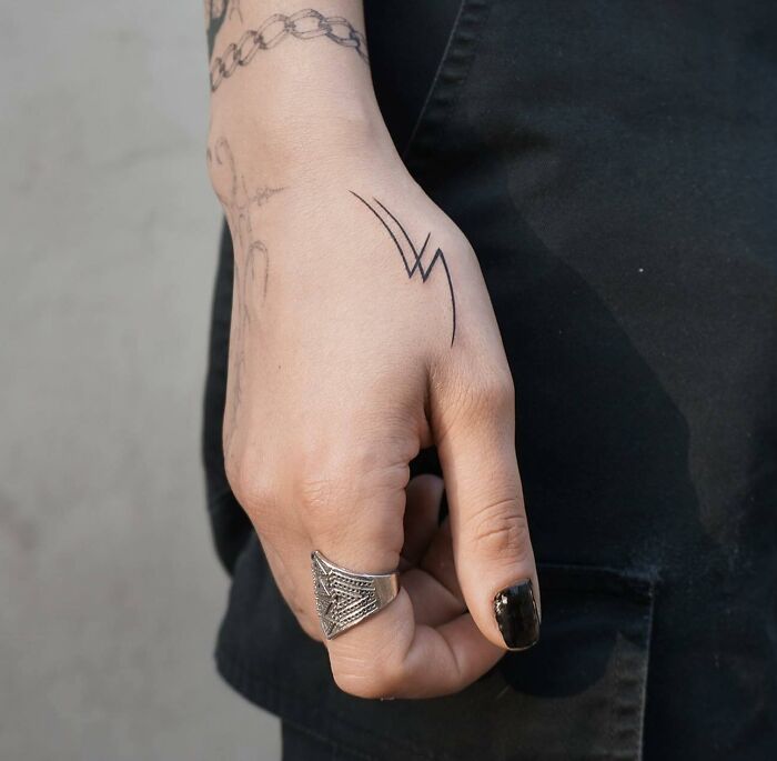 Close-up of a small hand tattoo featuring a simple lightning design, with a silver ring and black nail polish.