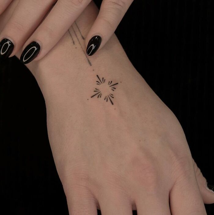 Minimalist small hand tattoo on wrist, black ink design with starburst pattern.