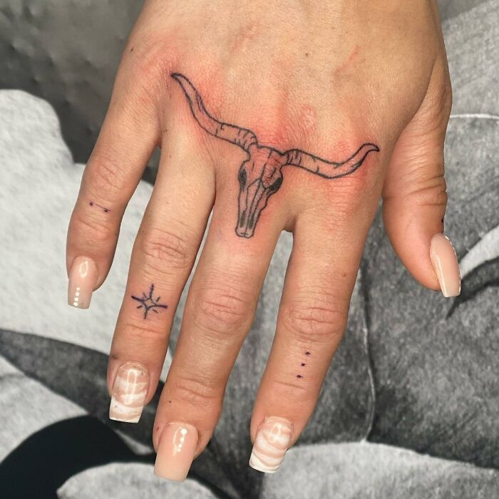 Small hand tattoo featuring a bull skull and star design on the fingers, showcasing intricate line work.