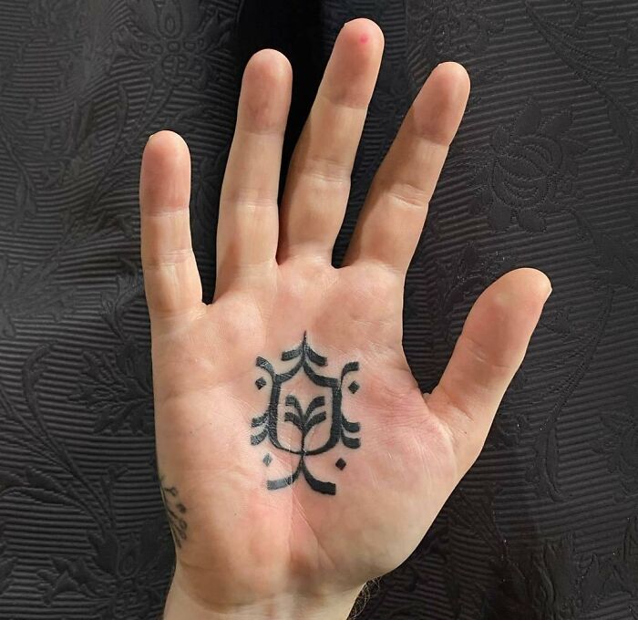 A small hand tattoo featuring a simple geometric pattern on the palm against a textured black background.