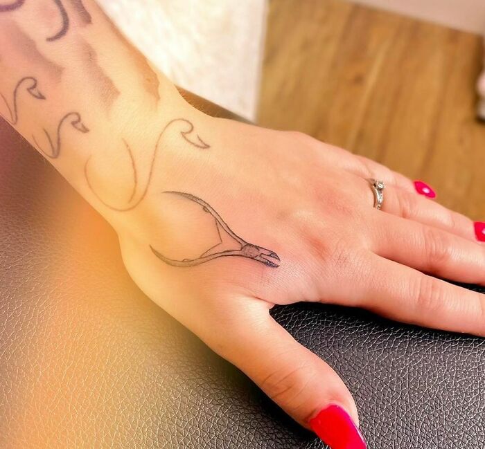Small hand tattoo of a wishbone on the back of a hand with red nails and a silver ring, showcasing delicate design.