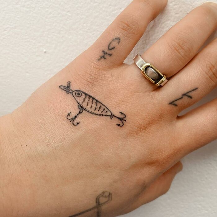 Small hand tattoo of a whimsical fish design with a ring and additional symbols on fingers.