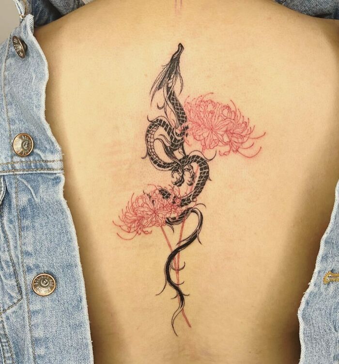 Intricate spine tattoo featuring a black dragon intertwined with red flowers on a person's back, beneath a denim jacket.