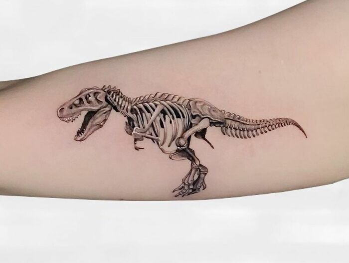 Detailed dinosaur tattoo featuring a T-rex skeleton on a person's forearm.