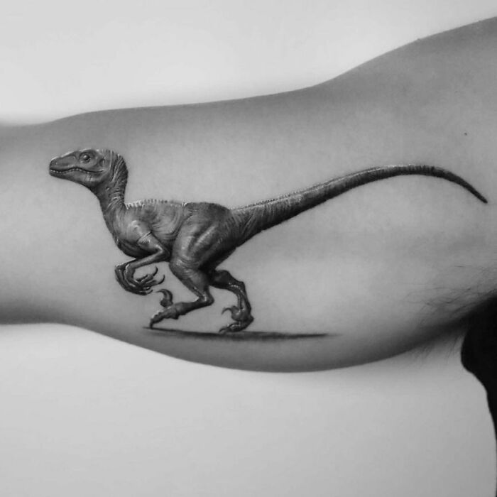 Cool dinosaur tattoo on an arm, showcasing a detailed velociraptor design in black and gray ink.