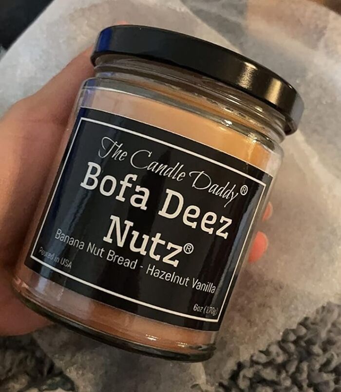 Hand holding a humorous scented candle labeled "Bofa Deez Nutz," featuring banana nut bread and hazelnut vanilla scent.