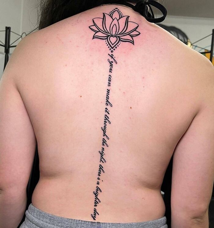 Back displaying a spine tattoo with a lotus design and inspirational script.