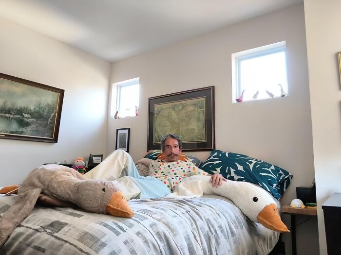 A man lounging on a bed with funny products, including a large stuffed goose, adding humor to his 2024.