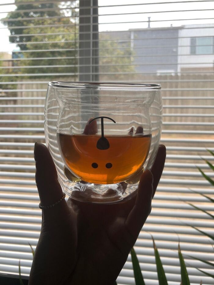 Hand holding a transparent glass mug with tea, featuring a cute face design. Perfect gift idea with Amazon Prime.