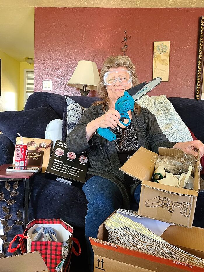 Woman holding a mini chainsaw gift, surrounded by opened presents on a couch.