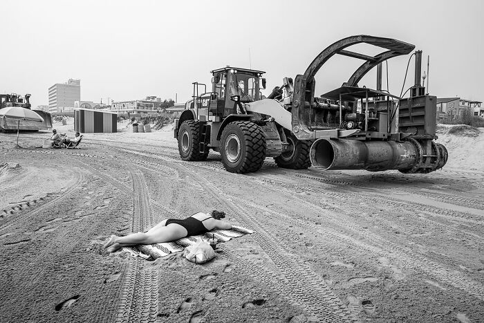 Black and white urban photography showing a person sunbathing on a beach near heavy machinery, capturing urban magic.