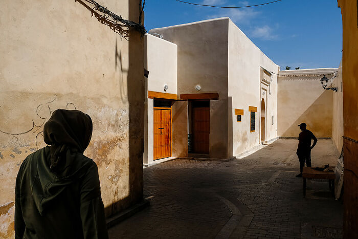 Urban street scene with contrasting light and shadow, featuring two figures and traditional building architecture.