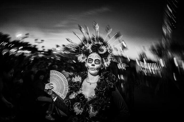 Black and white photo of a person in urban setting wearing Day of the Dead makeup and floral headpiece, capturing urban magic.