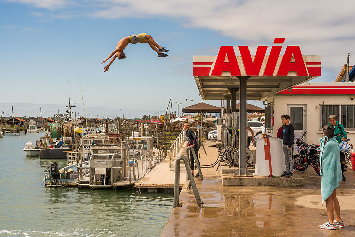 Young man diving into water near a gas station, capturing urban magic in a vibrant city scene for photography awards.