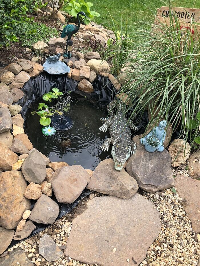 Garden pond with funny crocodile and heron sculptures, surrounded by rocks and greenery.