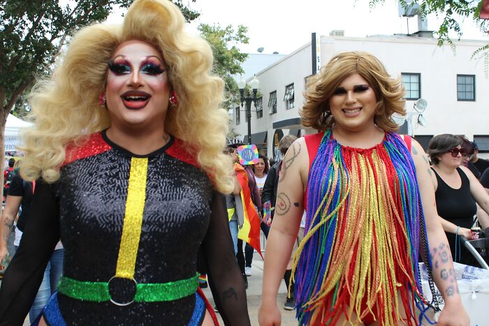 Two drag performers in colorful costumes smiling at an outdoor event, showing the world isn't a total nightmare.