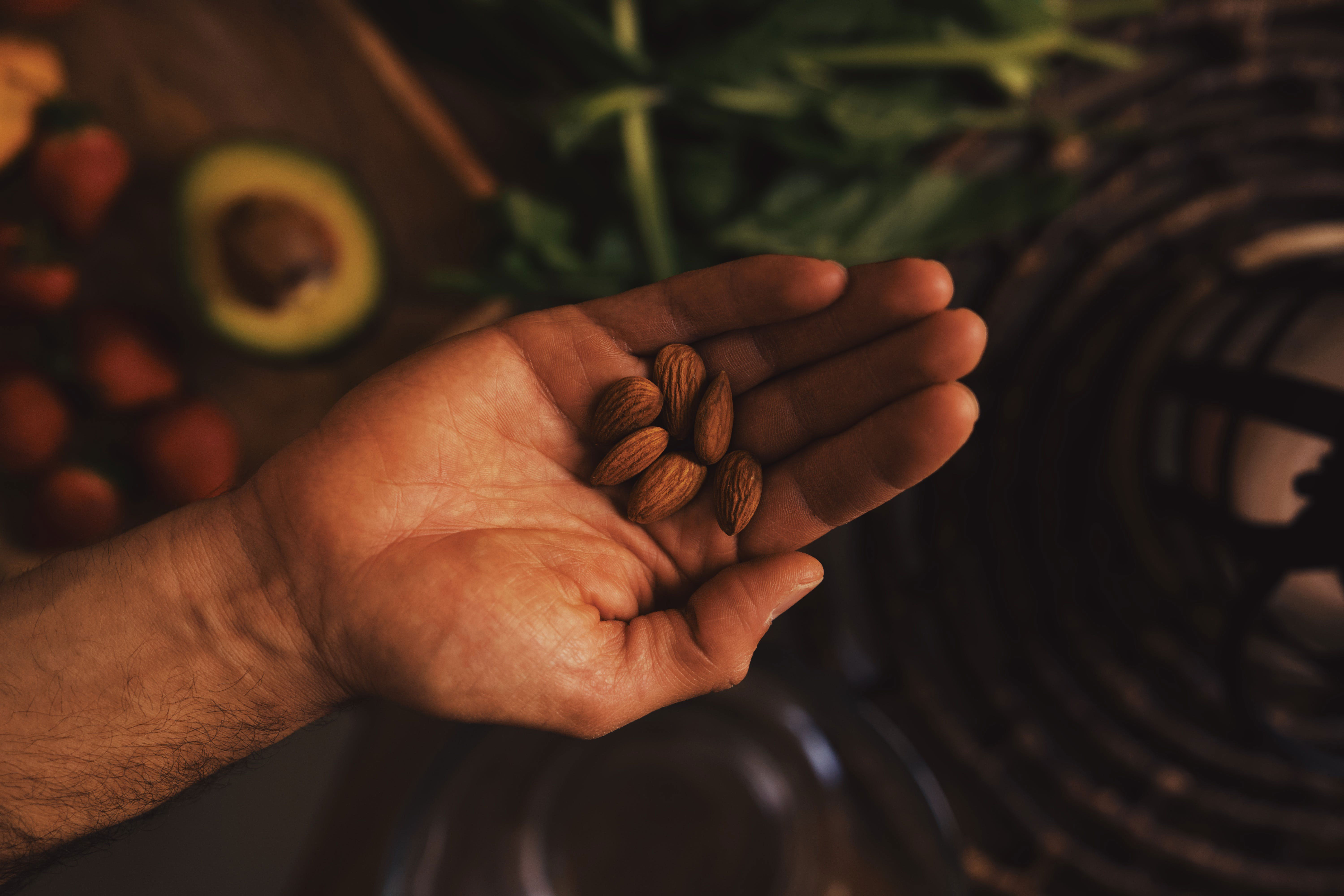 Hand holding almonds with leafy greens and fruits in the background.