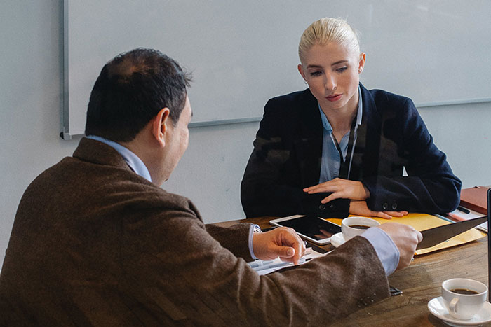 Two colleagues in a business meeting discussing normal workplace practices at a table with coffee cups and documents.