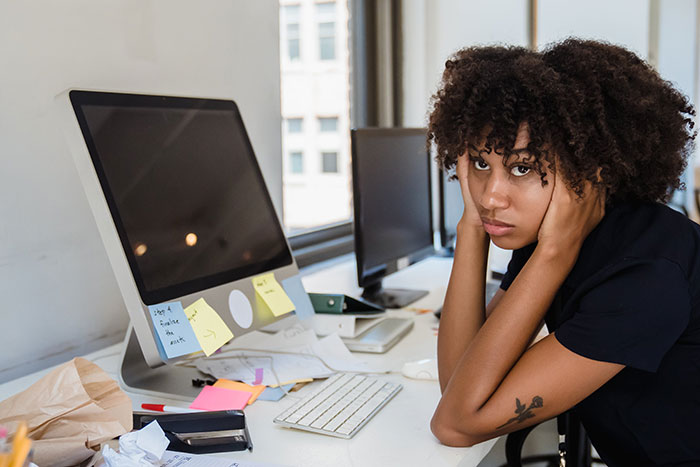 Frustrated young woman at cluttered desk illustrating stressful normal workplace practices people want to disappear.