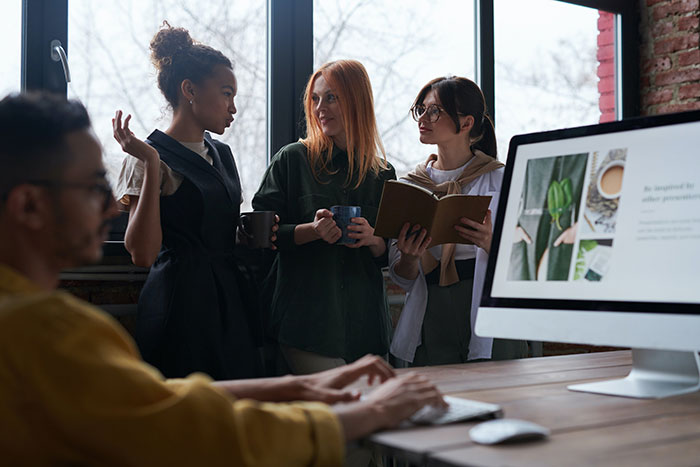 Three coworkers having coffee and discussing normal workplace practices while a colleague types on a computer nearby.