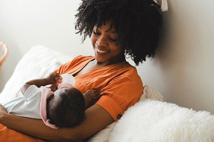 Woman in an orange shirt feeding a baby with a bottle, highlighting workplace practices related to work-life balance challenges.