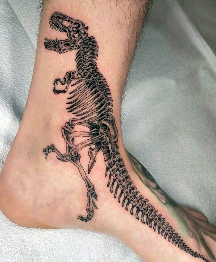 Dinosaur tattoo of a skeleton on a person's ankle, showcasing intricate design and detail.