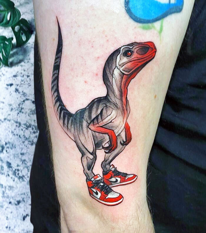Dinosaur tattoo featuring a raptor wearing red sneakers, showcasing creative tattoo artistry on an arm.