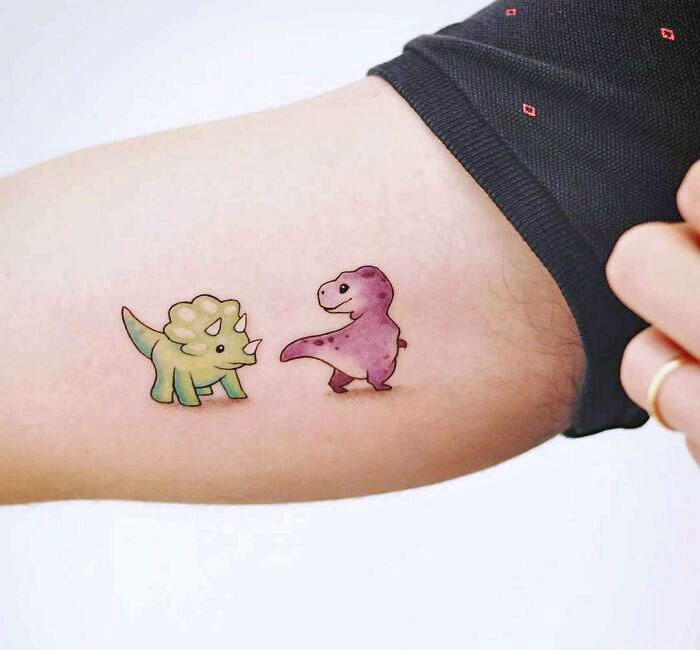 Cute dinosaur tattoo featuring a green triceratops and purple T-rex on a person's arm.