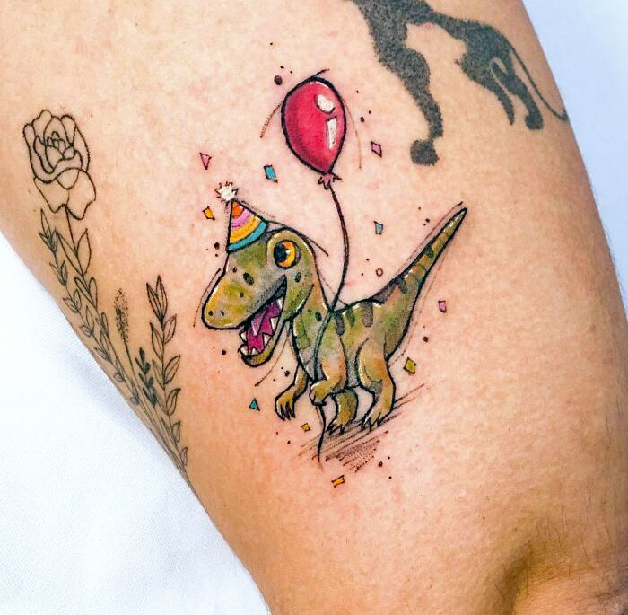 Colorful dinosaur tattoo with party hat and balloon, executed perfectly on skin.