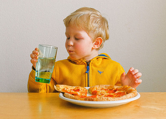 Young boy in a yellow hoodie holding a glass of water while sitting at a table with pizza, illustrating outdated parenting myths.