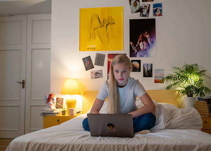 Teen girl sitting on bed using laptop in cozy bedroom, illustrating outdated parenting myths in a modern setting.