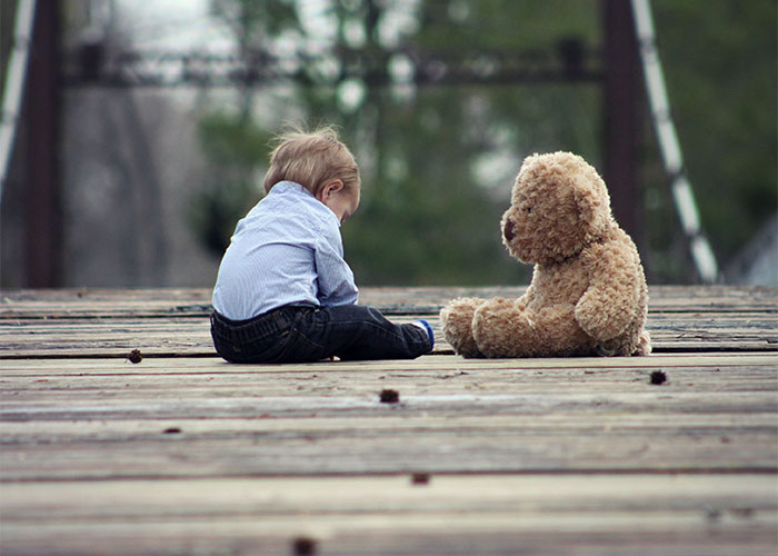 Toddler sitting on wooden bridge facing large teddy bear, illustrating outdated parenting myths in a natural outdoor setting.