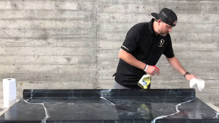 Man cleaning a black epoxy countertop with a spray bottle and cloth against a concrete wall background.