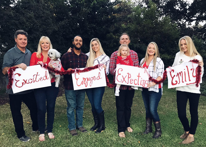 Family photo in a park with members holding signs: "Excited," "Engaged," "Expecting," "Emily," showcasing humorous family moments.